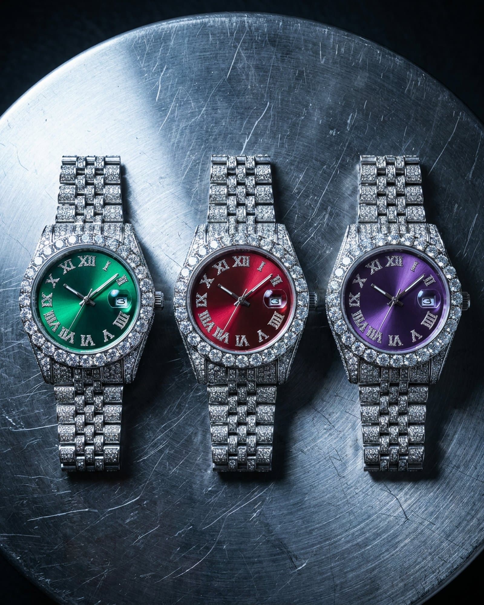Three iced round watches with green, red, and purple dials in a row on scratched steel
