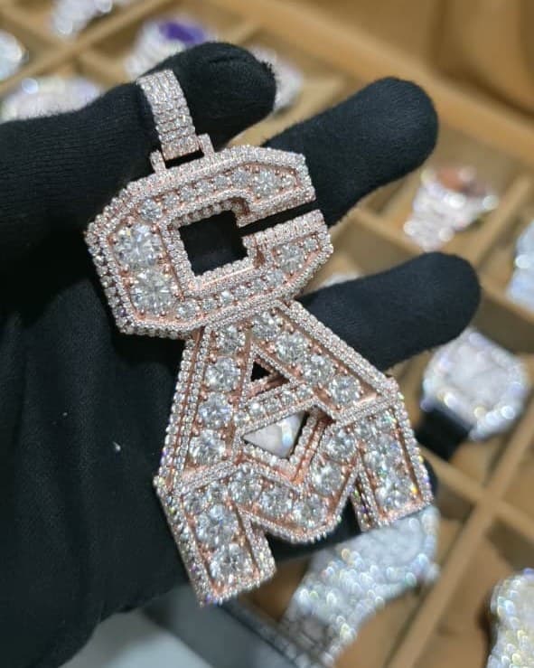 Rose gold pendant of three stacked block letters, large stones inside a pavé border, held in a black glove